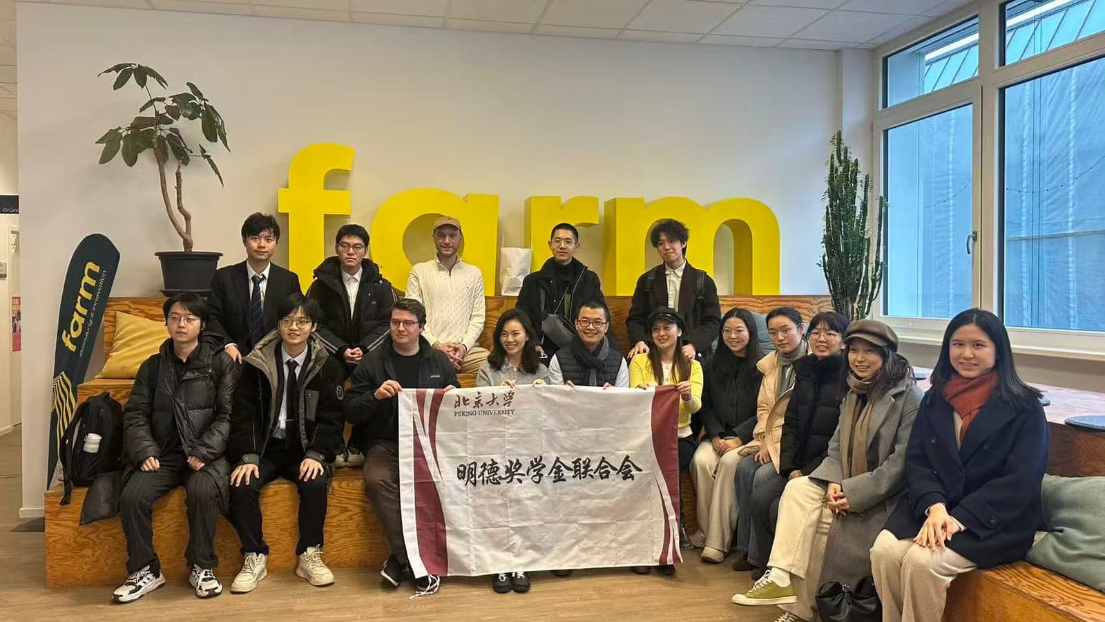 Group photo of a crowd of people holding a Peking University banner in a bright room, with a large yellow 'farm' sign on the wall behind them.
